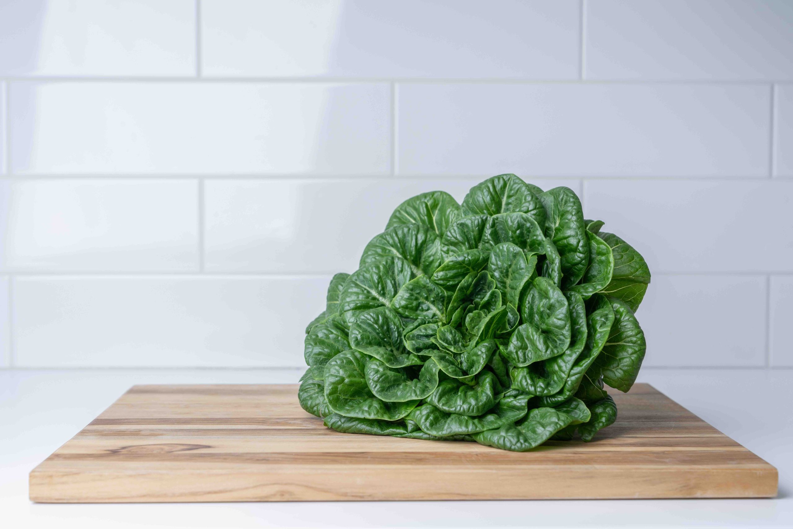 A head of Roma Romaine lettuce – rich green leaves – sits on a wooden cutting board, in a kitchen. The counter and tiled backsplash are off-white.